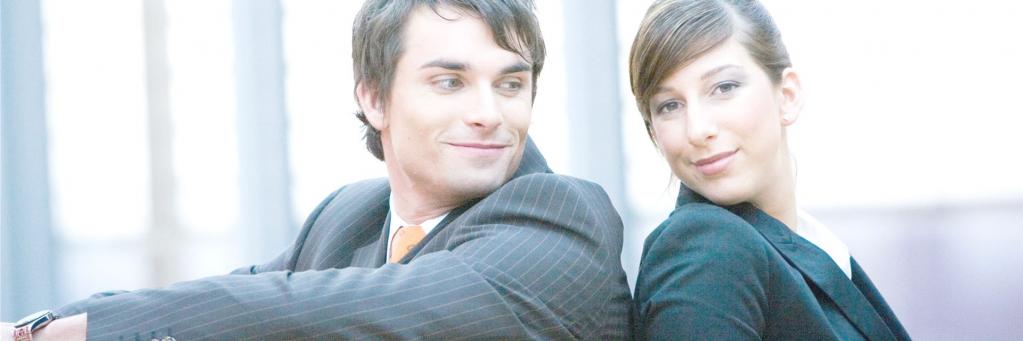 Ein Mann und eine Frau in Business-Kleidung lehnen Rücken an Rücken und blicken in die Kamera