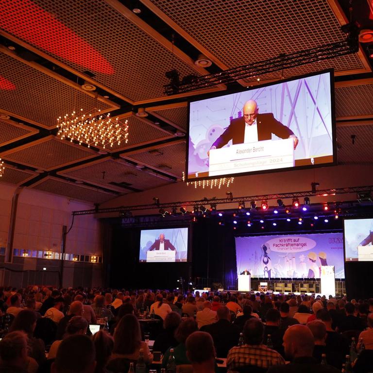 Blick zur Decke der Halle 1 der Westfalenhallen. Zu sehen sind mehrere Leinwände die mit projizierten Bildern vom Podium der Veranstaltung belichtet sind